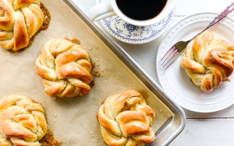 Nygräddade kardemummabullar på plåt med kaffe och en bulle på tallrik